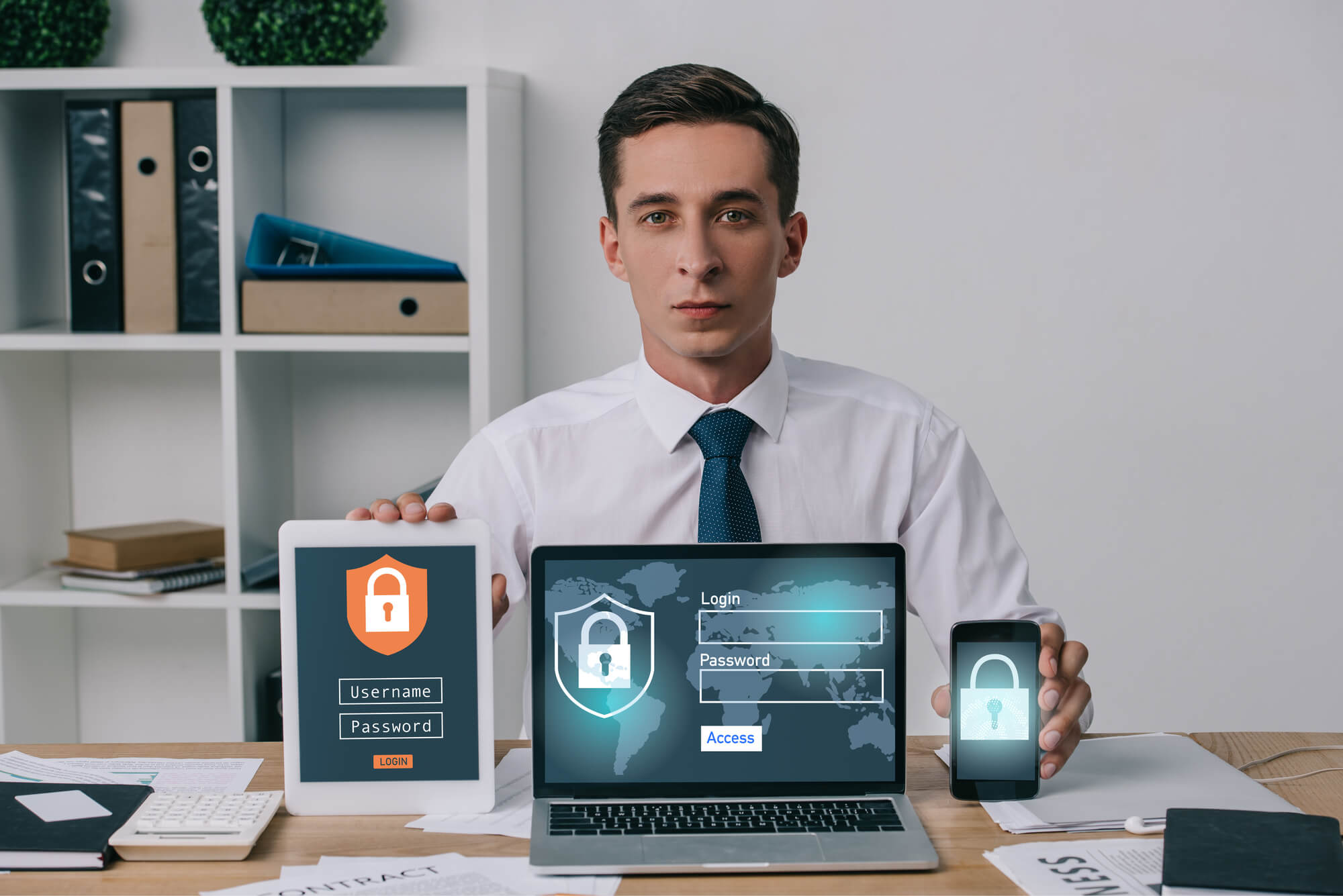 Portrait of businessman showing laptop, tablet and smartphone with cyber security signs on screens at workplace in office