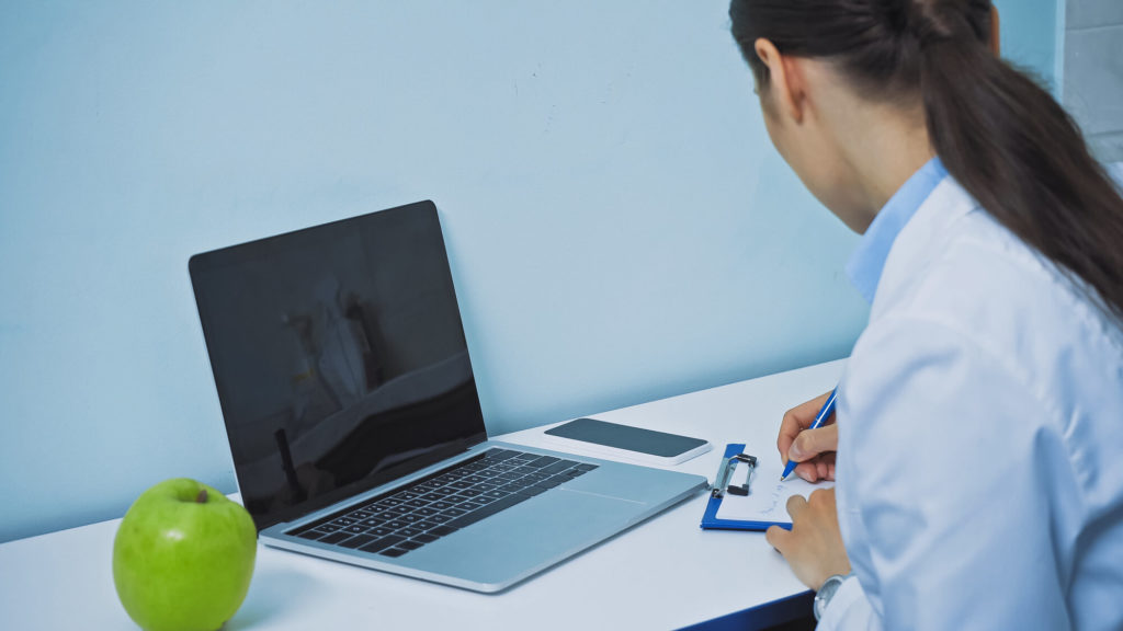 Dentist writing prescription on clipboard near laptop with blank screen and fresh apple